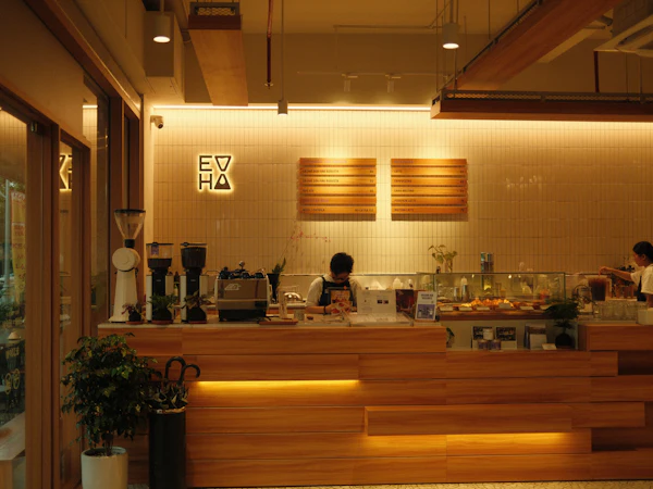 A long wooden coffee-shop counter lit from beneath, with grinders, an espresso machine and a menu board behind it.