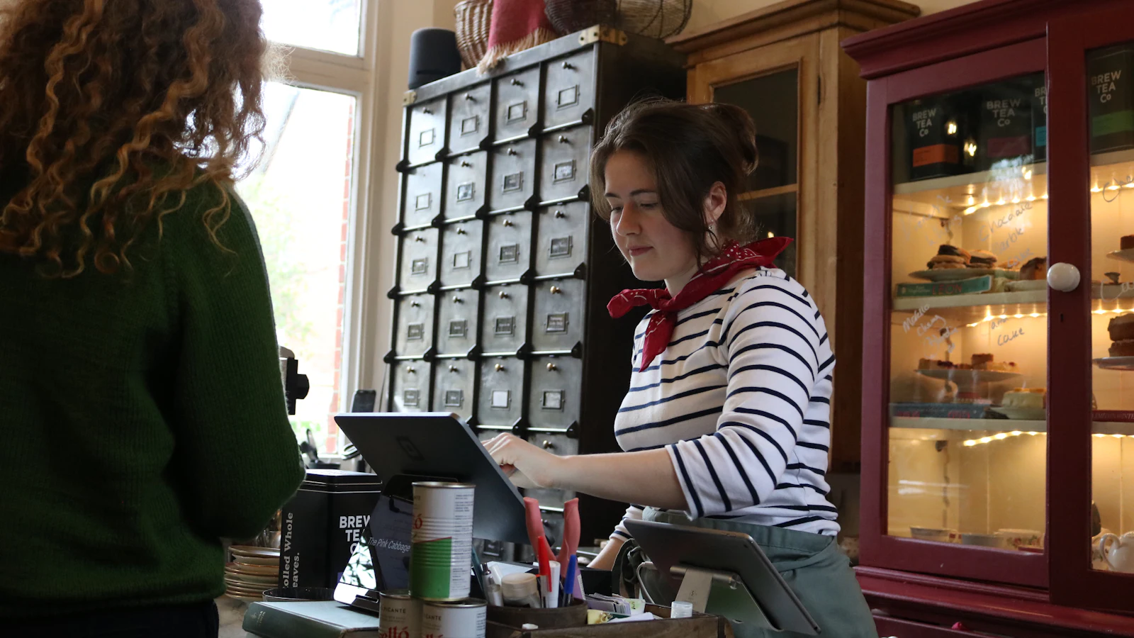 A member of staff at an independent café counter serving a customer, with a tablet till beside the pastry cabinet.