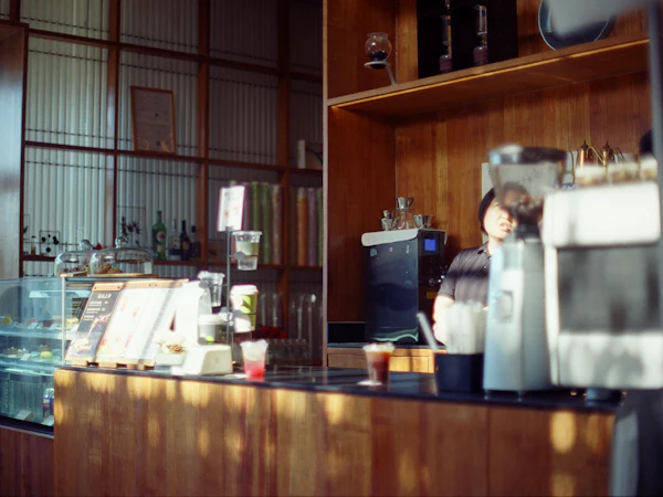 A sunlit wooden service counter with a chilled pastry case, cups and a coffee machine, and a member of staff working behind it.