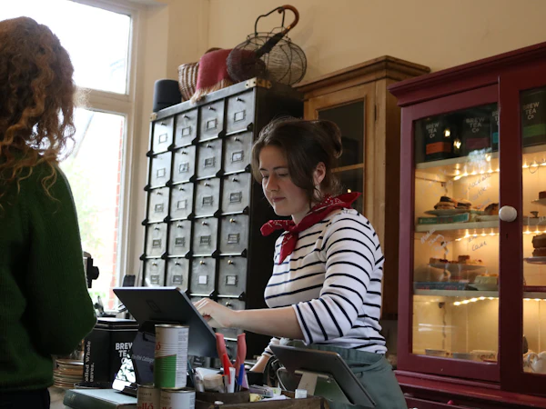 A member of staff at an independent café counter serving a customer, with a tablet till beside the pastry cabinet.